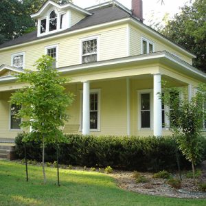 Two-story yellow house with front porch, bushes, and yard