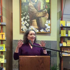 White woman in purple shirt speaking at lectern in bookstore with painting of man hugging a fish behind her