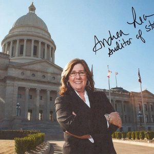 White woman with glasses and hoop earrings in suit smiling outside multistory building with large dome signed "Andrea Lea Auditor of State"