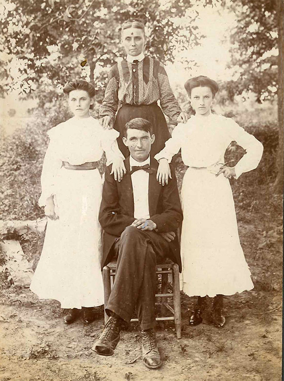 White man in suit and bow tie sitting with wife and daughters standing behind him