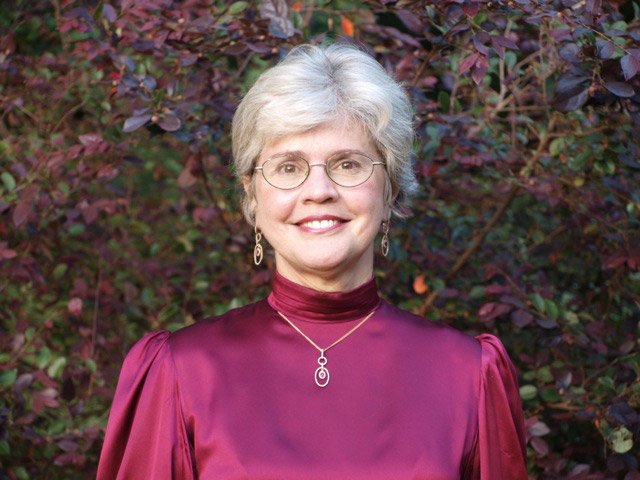 smiling white woman with gray hair wearing glasses earrings in red dress with necklace