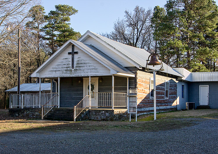 Single-story church building with covered porch log walls and bell in the front yard