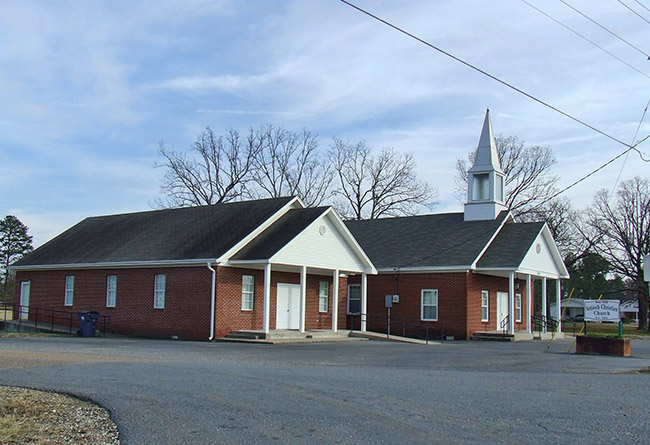 Single-story brick church buildings one with steeple and sign on parking lot