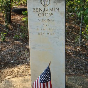 Weathered white gravestone with cross engraving above name and military ranks in forested area