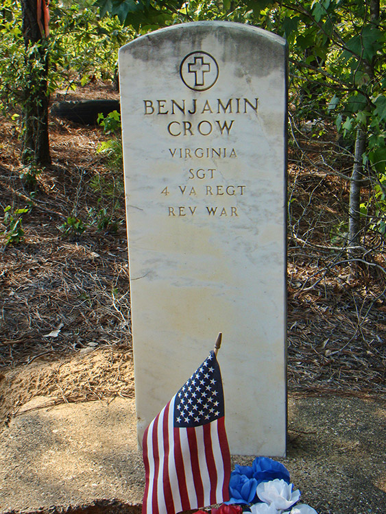 Weathered white gravestone with cross engraving above name and military ranks in forested area