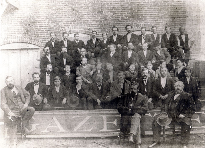 Group of white men in suits posing outside a brick building with large sign "Gazette" at their feet