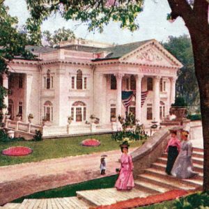 Two-story classical structure with four columns and American flag with in the foreground women in dresses on stairs in the shade of a tree