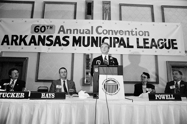 White men in suits seated at long table with white man in suit speaking at lectern with banner hanging behind them