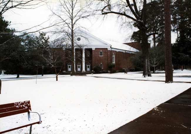 Snow covered quad and bench with crossing sidewalks leading to large multistory neoclassical brick building with white columns