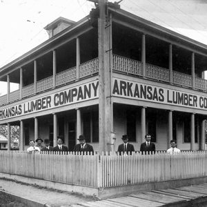 White men in suits standing inside fence with multistory building with covered porch and balcony behind them with large signs saying "Arkansas Lumber Company"
