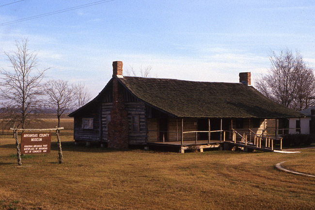 dogtrot-style log cabin with wooden front porch and ramp in vast open field and sign reading "Arkansas County Museum"