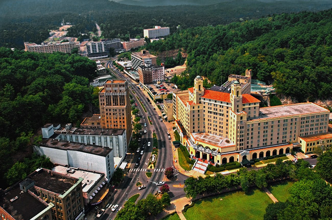 aerial view of downtown Hot Springs featuring the large somewhat triangular shaped Arlington hotel and various other tall buildings and boulevard with cars amid tree-covered hills