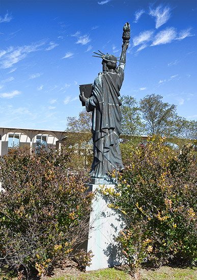 Reverse side of statue of woman holding torch in right hand and tablet in left hand on stone pedestal between two bushes