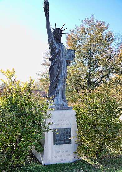 Statue of woman in robes and crown with torch in her right hand and tablet in her left on stone pedestal with plaque on it between two bushes