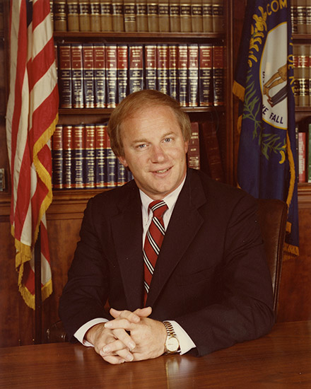 White man in suit and tie sitting at desk with flags and book shelves behind him