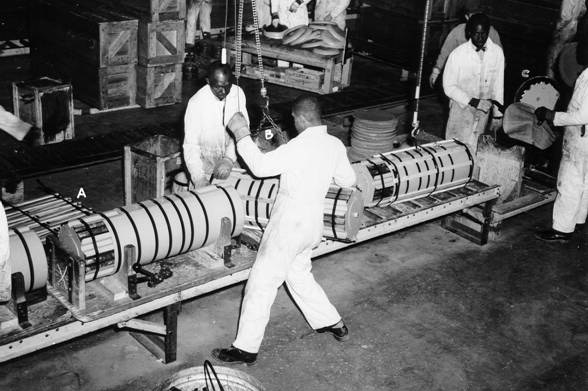 Black men factory workers in white coveralls at assembly line handling cylindrical objects on cranes