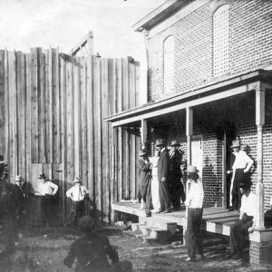 Group of white men in hats outside two-story brick building with barred windows and a scaffolding above which the top of a gallows can be viewed