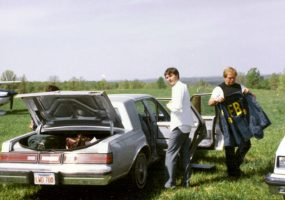 Three white men in field by sedans trunk open changing FBI uniform near small airplane