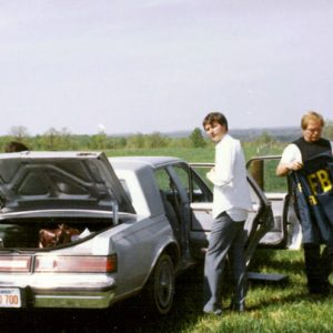 Three white men in field by sedans trunk open changing FBI uniform near small airplane