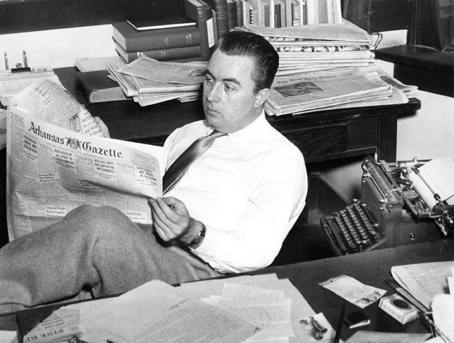 White man sitting at his desk reading a newspaper with typewriter behind him and stacks of newspapers around