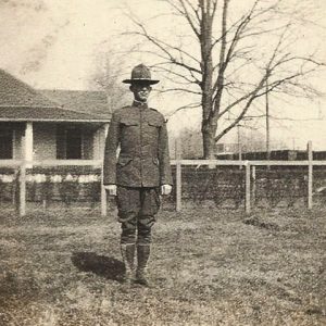 White man standing in military uniform with house fence and tree behind him