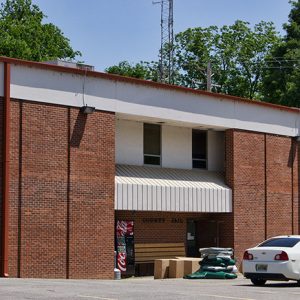 Two-story rectangular brick jail building with parking lot and coke machine boxes and parked car