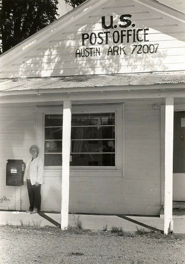 Old white woman standing by mailbox outside single-story post office building with covered porch