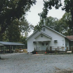 Single-story church building with steeple and covered porch on gravel parking lot with pavilion next to it
