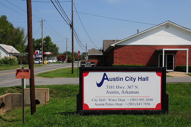 Street with "Austin City Hall" sign and brick building with gas station in the background