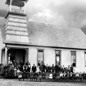 Crowd of white men women standing and white children sitting outside single-story church building with bell tower