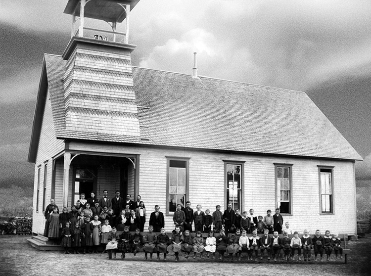 Crowd of white men women standing and white children sitting outside single-story church building with bell tower