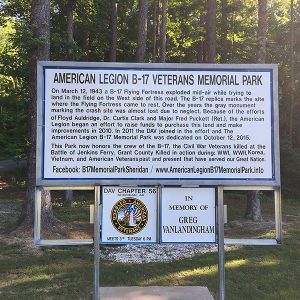 "American Legion B-17 Veterans Memorial Park" sign with text and smaller signs on grass with trees behind them