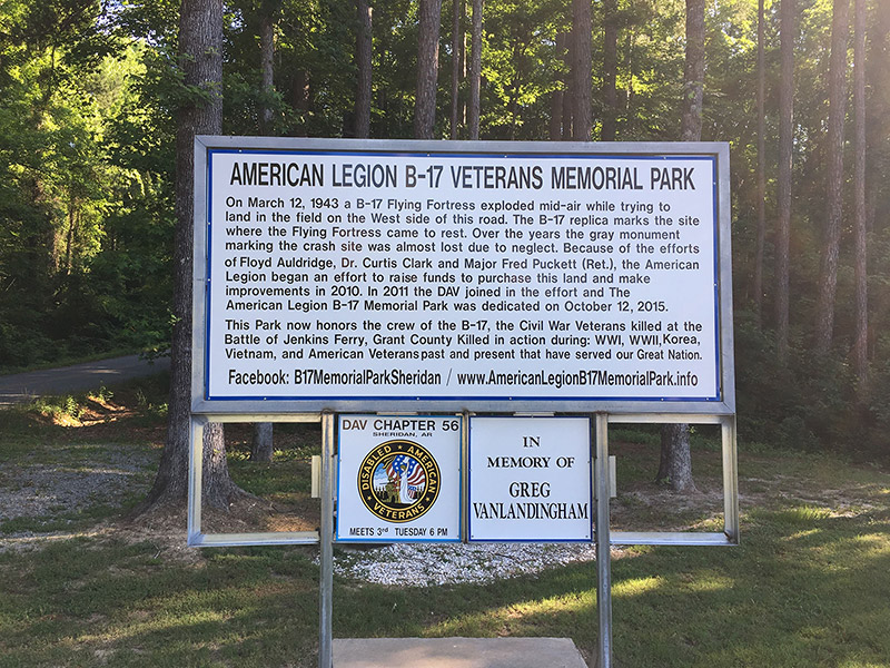 "American Legion B-17 Veterans Memorial Park" sign with text and smaller signs on grass with trees behind them
