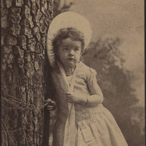 Young white girl with bonnet and dress standing next to a tree