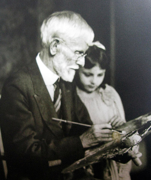 Old white man in suit and tie at easel with young white girl next to him