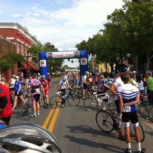 Crowd of people with bicycles wearing helmets and pads standing in city street at finish line with blue gateway in the background