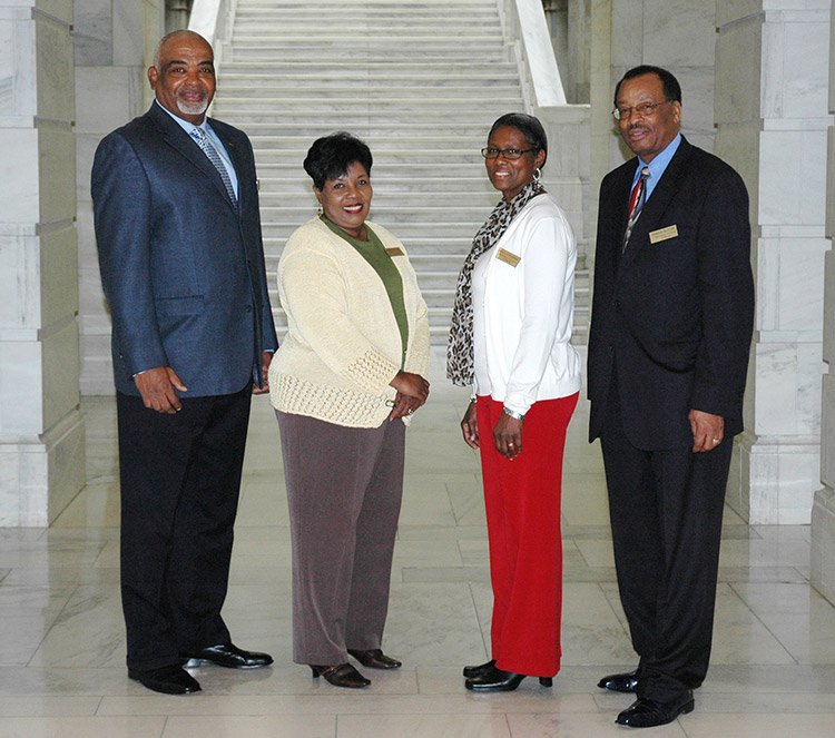 African-American men in suits and African-American women in white sweaters smiling in hallway with stairs behind them