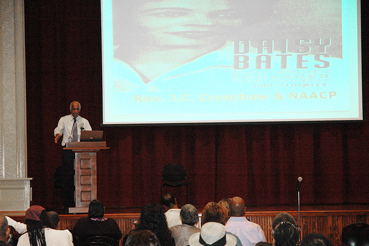 African-American man with gray hair speaking at lectern on stage with screen behind him