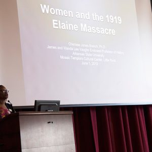 African-American woman speaking at lectern with presentation on large screen behind her
