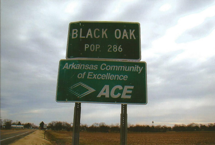 Weathered green "Black Oak" road sign on two-lane highway next to field