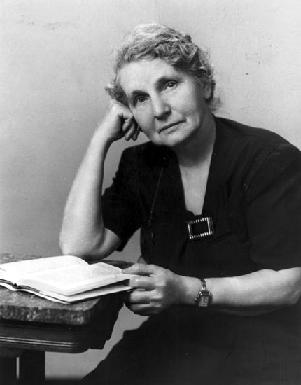 Older white woman in black posing with an open book on a table