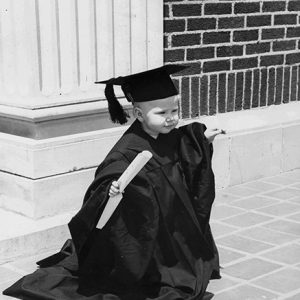 White toddler in graduation robes standing in front of brick wall