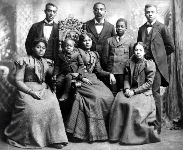 African-American women in formal dresses with a child sitting in chairs while men in suits stand behind them
