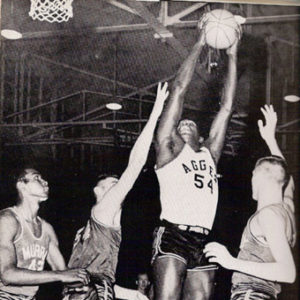 four men in basketball uniforms in mid game play with one African American player jumping in the attempt  to make a basket