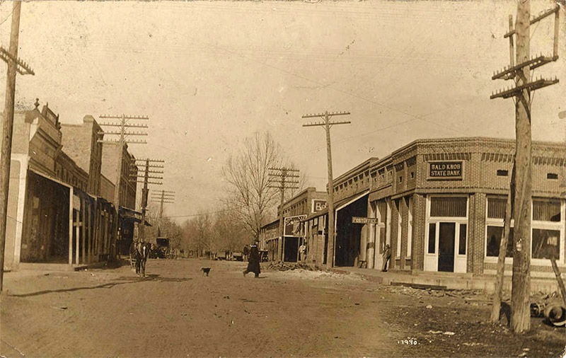 People walking on dirt road with brick buildings and telephone lines on both sides
