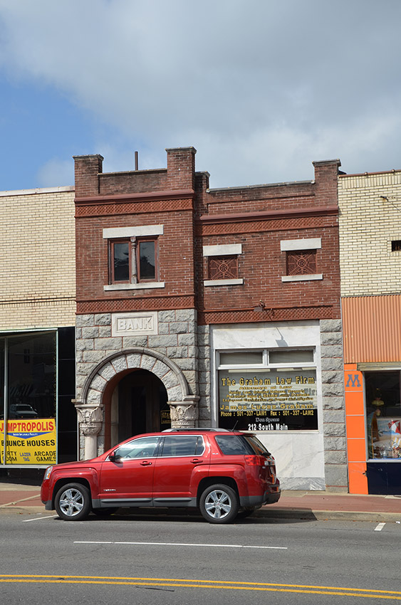 Two-story brick bank and storefront buildings on street with red SUV parked in front of them