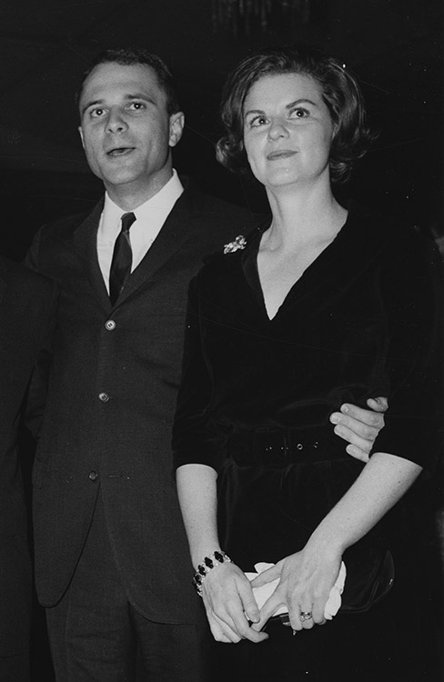 White man in suit standing with his arm around white woman's waist