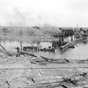 Steel truss bridge under construction over river with town buildings in the background