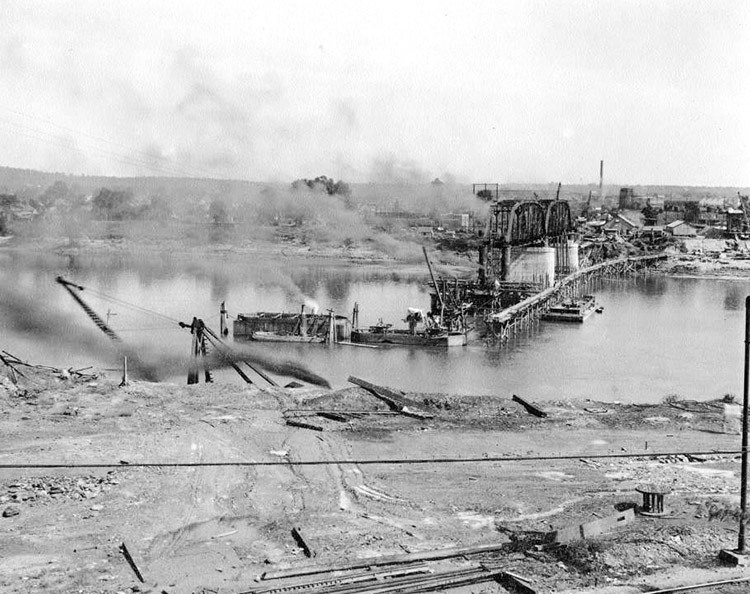 Steel truss bridge under construction over river with town buildings in the background