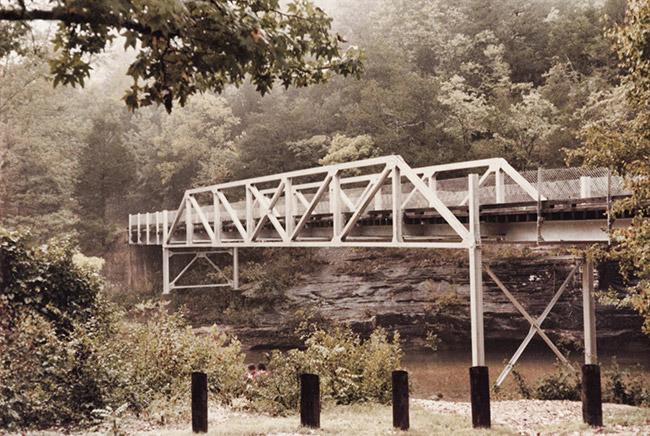 Steel truss foot bridge over creek with parking barriers in the foreground